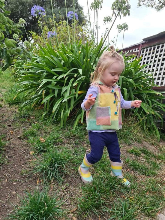 Young child wears a Pippa Studios best made from a recycled wool blanket that is a pastel rainbow colour. The child has matching gumboots and is walking in her garden. 